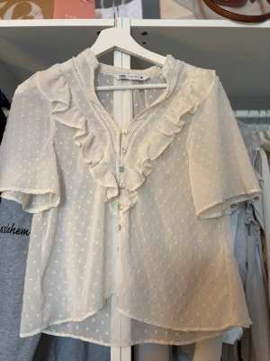 Vit blus med volanger från Zara - fint skick - Söt vit blus från Zara med korta ärmar och volangdetaljer längs knäppningen. Blusen är tillverkad i ett tunt, transparent tyg med små prickar och har dekorativa knappar framtill. (Andra bilden är inte min!)