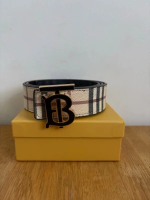 Rutig Burberry bälte med B-spänne - Snyggt bälte från Burberry med klassiskt rutigt mönster i beige, svart, vitt och rött. Bältet har ett stort metallspänne med Burberrys ikoniska B-logga. Perfekt accessoar för att lyfta din outfit.