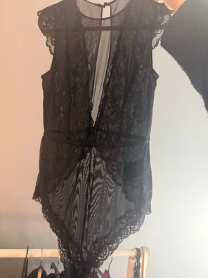 Svart spetsbody med meshdetaljer - Säljer en jättefin svart body i transparent mesh med vackra spetsdetaljer och djup v-ringning. Modellen är ärmlös med dekorativa kanter och knäpps i nacken. Perfekt för dig som vill ha något extra till din outfit.