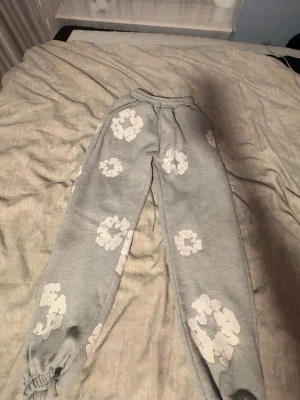 Grå mjukisbyxor med vita blommor - Snygga grå mjukisbyxor med vita blommönster över hela byxorna. Byxorna har resår i midjan och vid bensluten för en skön och avslappnad passform. Perfekta för chill dagar hemma eller när du vill ha en casual look.