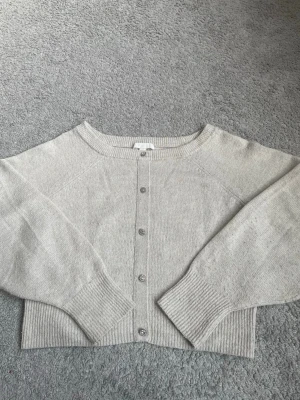 Beige stickad tröja med knappar - Snygg beige stickad tröja med rund halsringning och ribbade muddar. Koftan har raka ärmar och fem ”fejk” knappar framtill. Perfekt för en chill look.