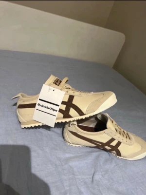 Onitsuka Tiger sneakers - Snygga beige sneakers från Onitsuka Tiger med bruna detaljer och klassiska ränder på sidan. Skorna har lågt skaft, snörning och ovandel i mocka och textil. Perfekta för dig som gillar retrostil och vill ha något unikt till din outfit.
