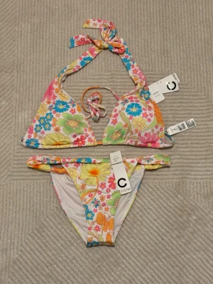 Färgglad blommig bikini från Cubus - Säljer en superfin bikini från Cubus med färgglatt blommigt mönster i rosa, gul, grön, blå och orange. Överdelen har knyt i nacken och ryggen, och bikinitrosan har låg midja. Perfekt för strandhäng och poolparty!