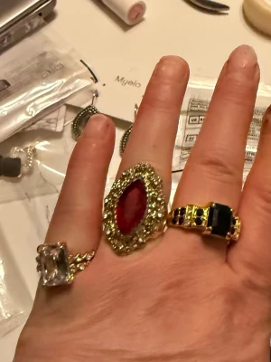 Tre statementringar med stenar - Säljer tre unika statementringar i guldtonad metall. En ring har en stor röd sten omgiven av små klara stenar, en har en stor rektangulär klar sten och den tredje har svarta stenar i en bredare design. Perfekta för dig som gillar att sticka ut.