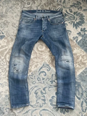 Blå slitna jeans från Jack & Jones - [FRI FRAKT 🚚]Säljer ett par blå jeans från Jack & Jones med slitningar och hål på knäna. Jeansen har fem fickor, smal passform och klassisk knappgylf. Snygg tvättad look och detaljerade slitningar för en trendig stil.
