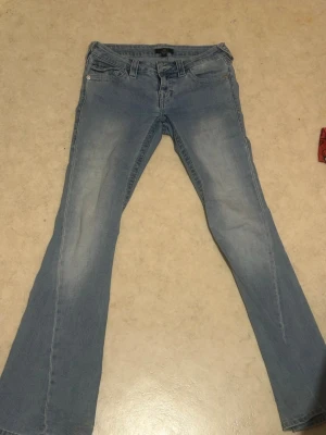 Blå bootcut jeans från True Religion  - Säljer ett par ljusblå bootcut jeans från True Religion med snygga slitningar framtill och detaljerade bakfickor med knappar. Klassisk femficksmodell och låg midja. Perfekta för dig som gillar retrovibbar och vill ha ett par jeans med unik look.