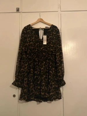 Leopardmönstrad kortklänning - Leopardmönstrad kortklänning från Neo Noir, modell Sima Leo Dress. Klänningen har långa ärmar med volang vid ärmslut och v-ringning med knyt i nacken. Sitter mer som en M och inte en L. Aldrig använd!