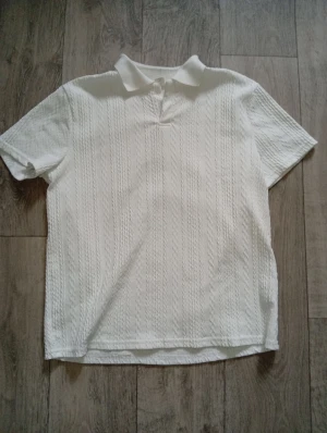 Vit kortärmad henley med struktur - Säljer en vit kortärmad henley med krage och subtilt strukturerat mönster. Skjortan har en klassisk passform och är tillverkad i ett mjukt material som känns skönt mot huden. Perfekt för dig som gillar stilrena och enkla plagg.