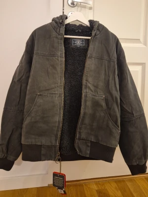 Grå bomberjacka från G.H. Bass & Co. - Snygg grå bomberjacka från G.H. Bass & Co. med huva och mjukt teddyfoder. Jackan har två stora framfickor, ribbade muddar vid ärmslut och nederkant samt dragkedja i metall. Perfekt för kyliga dagar och ger en avslappnad streetwear-look.