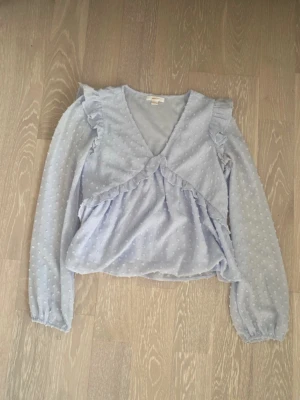 Ljusblå volangblus med prickar - Söt ljusblå blus med volangdetaljer och diskret prickmönster. Blusen har v-ringning, långa transparenta ärmar och resår vid ärmsluten. Materialet är tunt och ger en luftig känsla. Perfekt för dig som gillar romantisk och trendig stil.