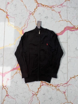 Mörk blå ziptröja från Polo Ralph Lauren - Kabelstickad mörk blå ziptröja från Polo Ralph Lauren med röd broderad logga på bröstet. Tröjan har ribbade muddar, hög krage och dragkedja framtill. Perfekt för en stilren och klassisk look.