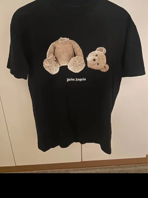Palm Angels svart t-shirt med björn - Svart t-shirt från Palm Angels med ikoniskt nalletryck på framsidan och stor Palm Angels-logga i vitt på ryggen. Klassisk passform och rund hals. Perfekt statement-plagg för dig som gillar streetwear. Storlek S och i nyckick. 