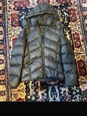 Svart pufferjacka med huva - Snygg svart pufferjacka från Gant med huva och dragkedja framtill. Jackan har två snedställda fickor med dragkedja och quiltad design i diagonalt mönster. Perfekt för kalla dagar och riktigt varm tack vare tjock vaddering.