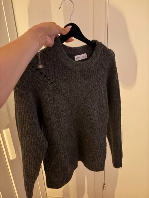 Mörkgrå stickad tröja från Lefties - Mysig mörkgrå stickad tröja från Lefties med rund halsringning och ribbade muddar. Tröjan har ett diskret mönsterstickat parti upptill och är perfekt för kyliga dagar. Enkel att matcha med jeans eller kjol.