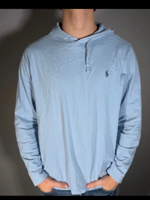 Ljusblå hoodie från Ralph Lauren - Ljusblå hoodie från Ralph Lauren med klassisk huva och lång ärm. Tillverkad i mjukt bomullsmaterial och har den ikoniska loggan broderad på bröstet. Enkel design med knappdetalj vid halsen, perfekt för en avslappnad och stilren look.