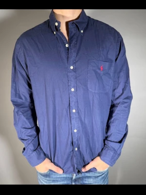 Blå skjorta från Ralph Lauren - Klassisk blå skjorta från Ralph Lauren med lång ärm, knappar framtill och bröstficka med röd broderad logga. Skjortan har en normal passform och är tillverkad i mjuk bomull. Perfekt för en stilren look.