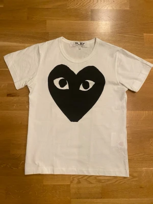 Vit Comme des Garçons PLAY t-shirt - Säljer en vit t-shirt från Comme des Garçons PLAY med det ikoniska svarta hjärta-ögon trycket på bröstet. Klassisk rund hals och korta ärmar. Tillverkad i mjuk bomull som är skön att bära. 