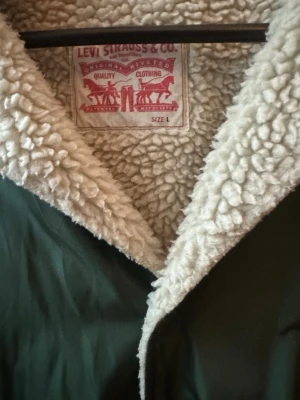 Grön teddyjacka från Levi's - Mörkgrön teddyjacka från Levi's med mjukt, vitt fuskpälsfoder och krage. Jackan har två fickor framtill och stängs med tryckknappar. Perfekt för kalla dagar och ger en avslappnad, trendig look.