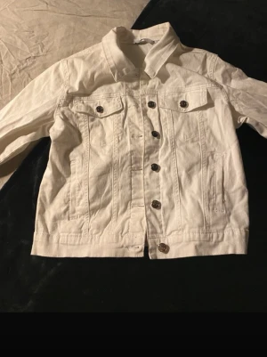 Vit jeansjacka med bruna knappar - Snygg vit jeansjacka med klassisk krage och knappar framtill. Två bröstfickor med lock och knapp samt två sidofickor. Rak passform och stilren design som passar till det mesta. Perfekt för dig som vill ha en fräsch och enkel look.