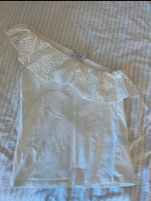 Vit oneshoulder topp - Söt vit oneshoulder topp från Holly & Whyte med volang och broderade spetsdetaljer längs axeln. Tillverkad i mjuk bomull, perfekt för sommarens alla tillfällen. Stilren och feminin design som sticker ut.