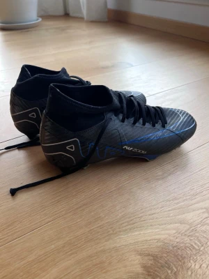 Nike Mercurial fotbollsskor svart/blå - Säljer ett par Nike Mercurial fotbollsskor i svart med blå detaljer och vita inslag. Skorna har hög krage för extra stöd runt ankeln och slitstarka dobbar för bra grepp på planen. Snygg, modern design med snörning och syntetmaterial för lätt känsla.