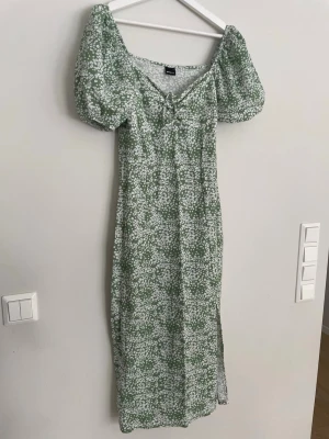 Grön blommig klänning Gina Tricot - Supersöt grön långklänning från Gina Tricot med vitt blommönster, puffärmar och snörning vid urringningen. Klänningen har en slits på sidan och är gjord i ett mjukt och bekvämt material, perfekt för sommarens alla tillfällen.