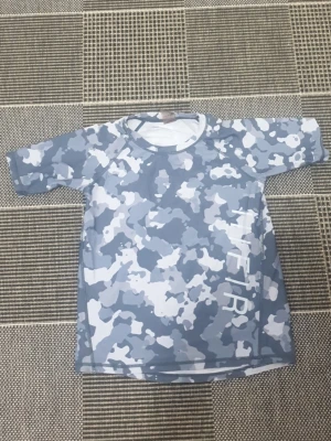 Blå camo strandtröja med tryck - Snygg strandtröja i blått och vitt camomönster med korta ärmar och rund hals. Materialet är snabbtorkande och perfekt för stranden. Tryck med texten 'THE IR' på sidan framtill. Perfekt för soliga dagar vid vattnet.