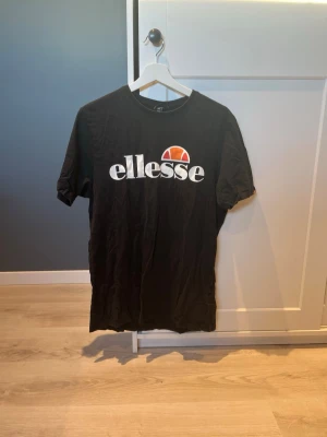 Svart Ellesse t-shirt med logga - Svart t-shirt från Ellesse med stor vit logga och orange detalj på bröstet. Klassisk rund hals och korta ärmar. Perfekt för en avslappnad och sportig stil.