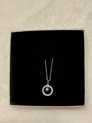 Silverhalsband med cirkelhänge - Snyggt halsband i silverfärg med ett cirkelformat hänge dekorerat med gnistrande stenar runt om och en större sten i mitten. Kedjan är tunn och elegant, perfekt för att lyfta vilken outfit som helst. Kommer i en stilren ask.