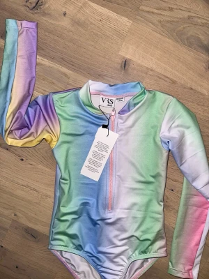 Regnbågsfärgad långärmad baddräkt VRS - Snygg långärmad baddräkt från VRS i regnbågsfärgat mönster med dragkedja framtill. Materialet är stretchigt och snabbtorkande, perfekt för strand eller pool. Färgerna går i pastelltoner av rosa, blått, gult, grönt och lila. Rund halsringning och figurnära passform.