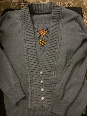 Grå kofta med pärldetaljer - Snygg grå kofta med ribbad krage och knappslå. Framsidan har dekorativa pärlor i orange, gult och grönt som bildar ett blommönster. Långa ärmar och mjukt stickat material gör den både bekväm och stilren. Perfekt för dig som vill ha något extra i garderoben.