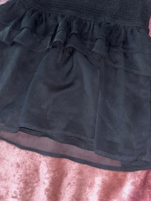 Svart volangkjol i mesh - Säljer en svart kortkjol med dubbla volanglager och transparent meshmaterial. Kjolen har en smockad midja som ger en bekväm passform och en luftig känsla. Perfekt att styla med en enkel topp för en trendig look. Funkar att använda som både en kjol med även en topp 