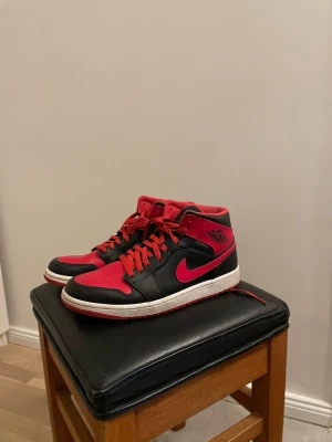Air Jordan 1 Mid Svart/Röd - Säljer ett par Air Jordan 1 Mid sneakers i svart och rött läder med vita sulor. Klassisk Nike Swoosh på sidan och Air Jordan-logga vid ankeln. Skorna har röda skosnören och perforerad tåbox för extra ventilation. Perfekta för dig som gillar streetwear och ikonisk design.