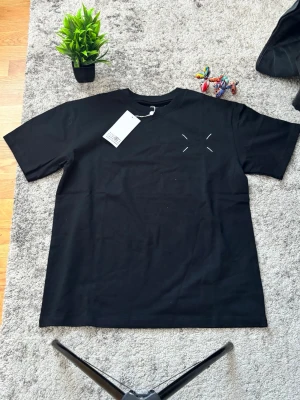 Maison Margiela svart t-shirt - Svart t-shirt från Maison Margiela med ikoniska vita sömmar fram och stort tryck med siffror och logga på ryggen. Klassisk passform och rund hals. Perfekt för dig som gillar streetstyle och vill ha något från ett high fashion-märke.