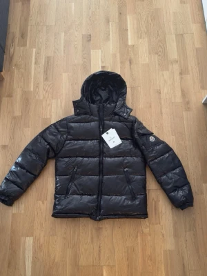 Svart dunjacka från Moncler med huva - Snygg svart dunjacka från Moncler med glansig finish och fast huva. Jackan har två dragkedjeförsedda fickor framtill och Moncler-logga på ärmen. Perfekt för kalla vinterdagar och riktigt stilren look.