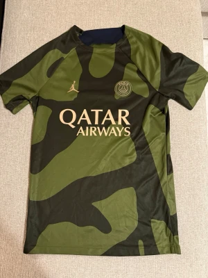 PSG x Jordan camo t-shirt grön - Säljer en grön och svart camo t-shirt från PSG x Jordan med Qatar Airways-tryck på bröstet. Tröjan har rund hals, korta ärmar och är tillverkad i ett lätt och snabbtorkande material. Perfekt för träning eller streetwear.