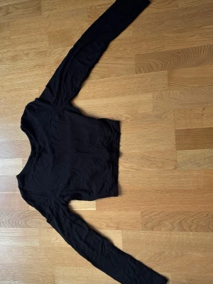 Svart croppad topp med knyt baktill - Säljer en svart croppad långärmad topp med knyt i midjan. Toppen har en enkel och stilren design med rund halsringning och är tillverkad i ett mjukt material som känns skönt mot huden. Säljer på grund av för liten i storlek och kommer inte till användning!