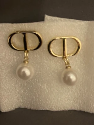 Mode örhängen  - Snygga örhängen från Dior med guldfärgad metall och hängande vita pärlor. Örhängena har den ikoniska CD-loggan upptill och en klassisk, elegant design som passar perfekt till att lyfta din stil.