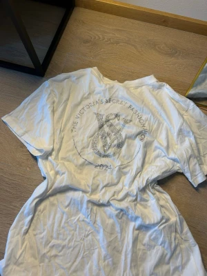 Vit t-shirt Victoria's Secret - Vit t-shirt från Victoria's Secret med glittrande VS-logga och text på ryggen samt en liten silverfärgad logga framtill. Klassisk rund hals och korta ärmar. Tillverkad i mjuk bomull för en bekväm känsla. #angelwings #fashionshow2024