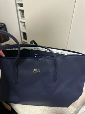 Mörkblå shoppingväska från Lacoste - Snygg mörkblå shoppingväska från Lacoste med klassisk krokodillogo framtill. Väskan har två långa handtag, dragkedja upptill och ett innerfack med dragkedja. Tillverkad i tåligt syntetmaterial med Lacoste-tryck på insidan. Perfekt för dig som vill ha både stil och funktion.