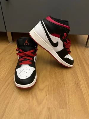 Air Jordan 1  - Snygga Air Jordan 1 Mid sneakers i svart, vitt och rött med klassisk Nike Swoosh på sidan. Skorna har röda skosnören, perforerad tåbox och ikoniska Jumpman-loggan på plösen. Ovandelen är i läder och textil för en clean och sportig look.