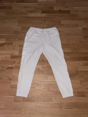Vita mjukisbyxor från Hugo Boss - Säljer ett par vita mjukisbyxor från Hugo Boss med resår i midjan och muddar vid bensluten. Byxorna har en stilren design med diskreta detaljer längs sidorna och är tillverkade i mjukt material som känns skönt mot huden. Perfekta för en avslappnad look.
