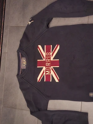 Mörkblå stickad tröja Morris - Snygg mörkblå stickad tröja från Morris med Union Jack-flagga och Morris-text broderat på bröstet. Tröjan har rund halsringning och ribbade muddar vid ärmslut och nederkant. Perfekt för dig som gillar klassisk brittisk stil.