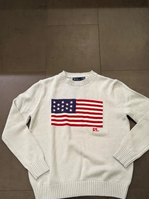 Vit stickad tröja Ralph Lauren - Vit stickad tröja från Ralph Lauren med amerikansk flagga i blått, rött och vitt på bröstet samt RL-broderi. Tröjan har rund halsringning, långa ärmar och ribbade muddar vid ärmslut och nederkant. Perfekt för en klassisk och avslappnad stil.