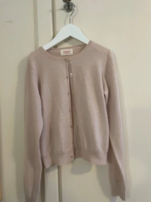 Ljusrosa kofta från Maan - Söt ljusrosa kofta från Maan med rund halsringning och knappar framtill. Koftan är ribbstickad vid ärmslut och nederkant, har en klassisk rak passform och är tillverkad i mjukt material. Perfekt för lager på lager och enkel att matcha.