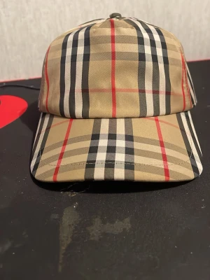 Burberry beige rutig keps - Säljer en klassisk Burberry keps i beige med det ikoniska rutmönstret i svart, vitt och rött. Kepsen har justerbar snapback och en patch med texten 'BURBERRY LONDON ENGLAND' bak. Perfekt för dig som vill ha en stilren och trendig accessoar.