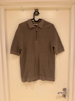 Beige stickad polotopp från Dobber - Säljer en beige stickad polotopp från Dobber med korta ärmar och ribbad struktur. Toppen har en klassisk krage och dragkedja framtill som ger en snygg detalj. Perfekt att styla med jeans eller kjol för en trendig look.                                                               Modellen är 185cm lång och väger 82kg.