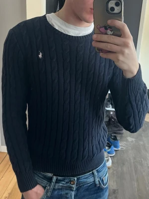 Mörkblå kabelstickad tröja Polo Ralph Lauren - Säljer en mörkblå kabelstickad tröja från Polo Ralph Lauren med den klassiska vita logon broderad på bröstet. Tröjan har rund halsringning, ribbade muddar och är tillverkad i mjukt material. Perfekt för dig som gillar stilren och tidlös look. Storlek M dam passar herr xs