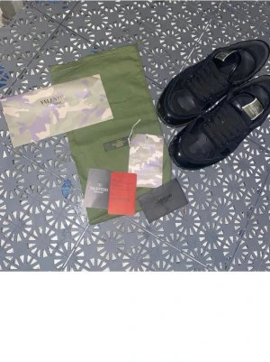 Svarta Valentino Garavani sneakers - Snygga svarta sneakers från Valentino Garavani med diskret kamouflagemönster på ovandelen. Skorna har en klassisk låg siluett, svarta skosnören och detaljer i läder. Perfekta för dig som vill ha en stilren men unik look.