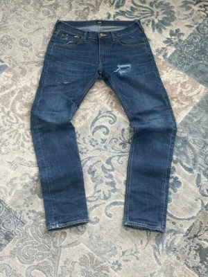 Mörkblå slitna jeans från Lee - [FRI FRAKT 🚚] Säljer ett par mörkblå jeans från Lee med slitningar och distressed detaljer på låret och fickan. Klassisk femficksmodell med raka ben och knappgylf. Jeansen har en cool vintagekänsla och passar perfekt till en avslappnad stil.